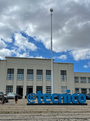 Photo of the building housing the Instituto Superior Técnico in Lisbon, Portugal. At front of building are steps and a sculptural sign in blue that says "@tecnico."