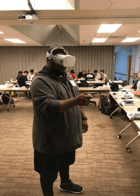 A person stands in a classroom using a virtual reality headset with people seated at tables in the background.
