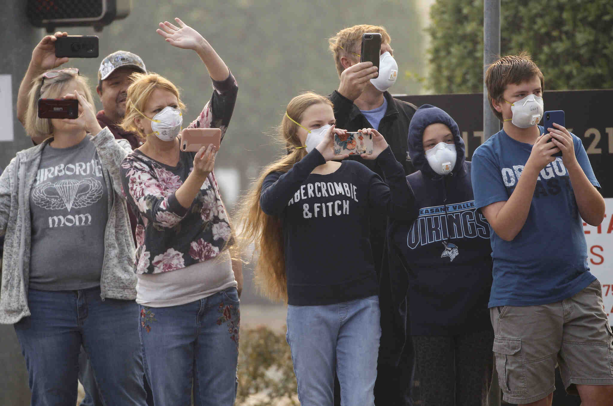 A group of people stand on a street wearing air masks and taking video on their cellphones.