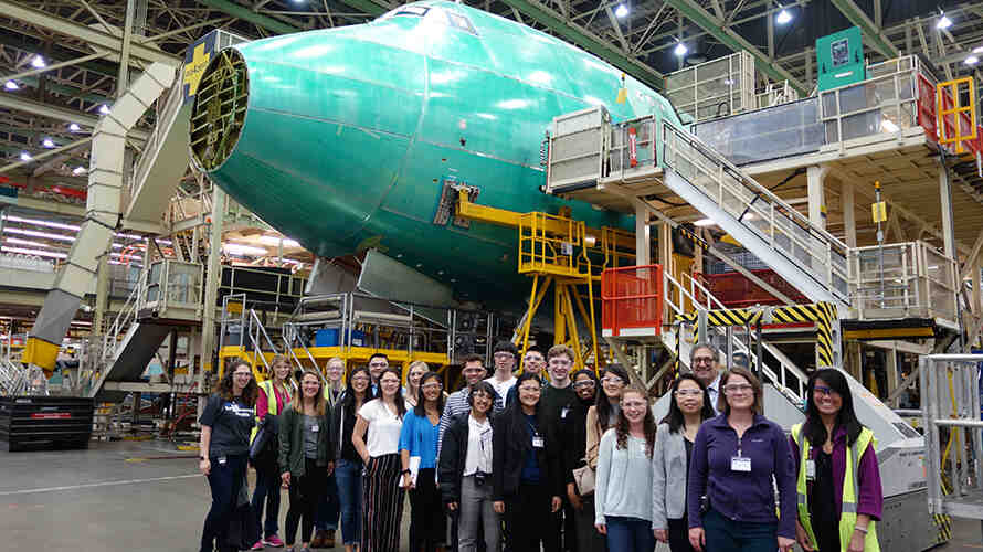 A group of DEOHS students, staff and faculty stand in front of an airplane under construction at a Boeing manufacturing facility.