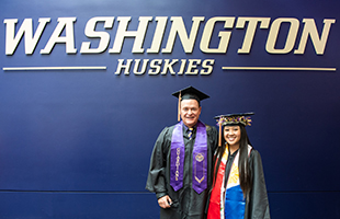 Castner and UW Senior Xamantha Curameng stand in graduation garb in front of a purple wall with light brown lettering that reads Washington Huskies.