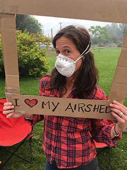 A volunteer in a red plaid shirt with a filtering mask over her face poses with a cardboard frame that reads "I love my airshed", greenery in the background.