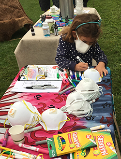 A little girl wearing an air filtering mask colors another mask with markers at a table.