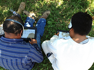 Two agricultural workers bent over tablets to take a survey while seated on grass.