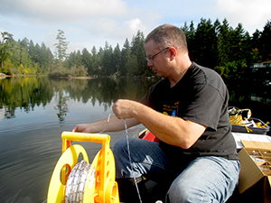 UW research technologist Corey King measures the water transparency of Lake Killarney.  He sits in a boat with the lake and trees int he background, working with a large spool of rope.  Photo: James Gawel.