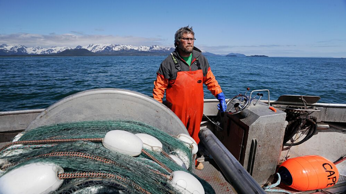 Fisherman stands on the deck of a silver, metal boat, mountains and the ocean behind him and fishing equipment around him.