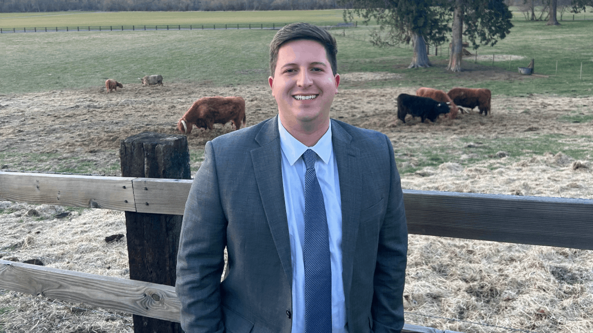 Jorge Rivera-Gonzalez stands in a suit and tie in front of a fence and pasture with cows.