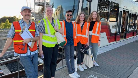Five people in safety vests stand in front of a light rail train.