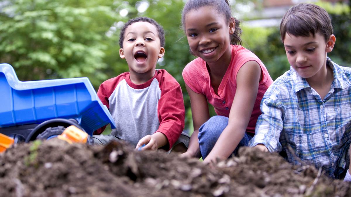 Three kids play outside in the dirt with a blue toy dumptruck. They look happy.  