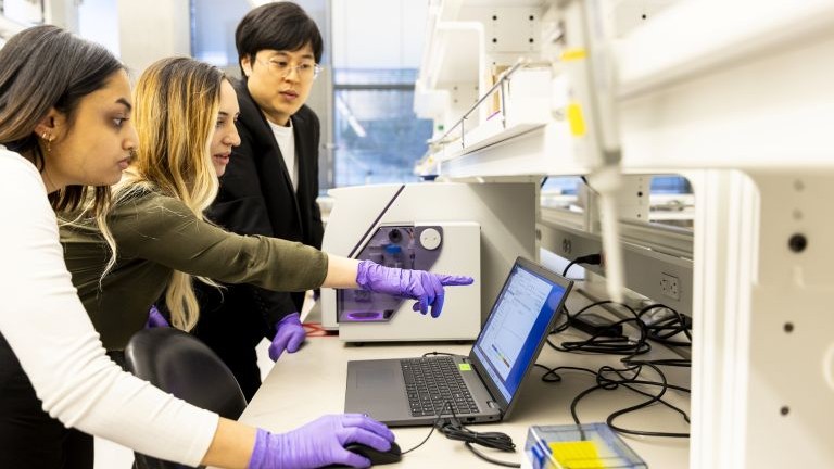 Three people work on a computer. One of them is wearing a lab coat and purple gloves.