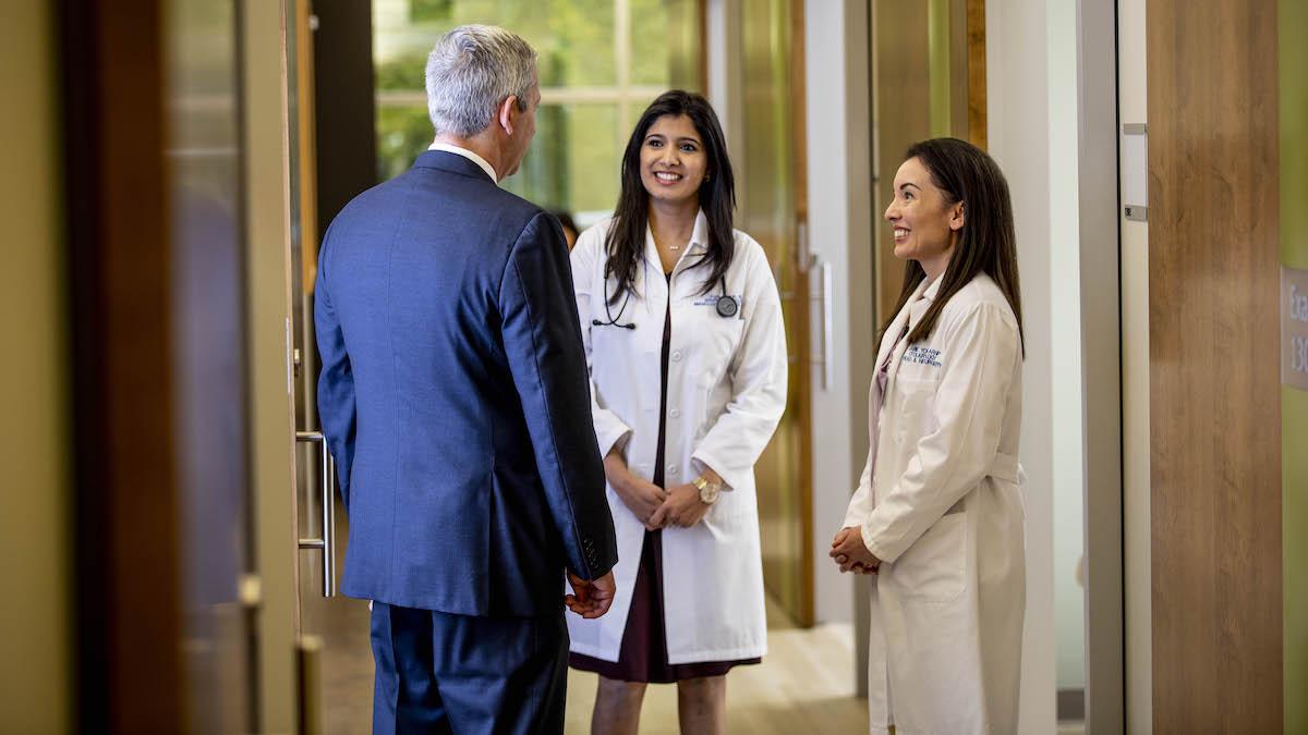 Two people in lab coats talk to a person in a blue suit. They are standing in the hallway of a hospital.