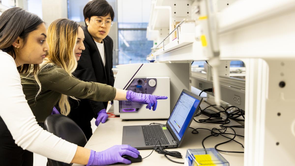 Three researchers look at a laptop screen. They are surrounded by lab equipment. One of the researchers is pointing at the screen.