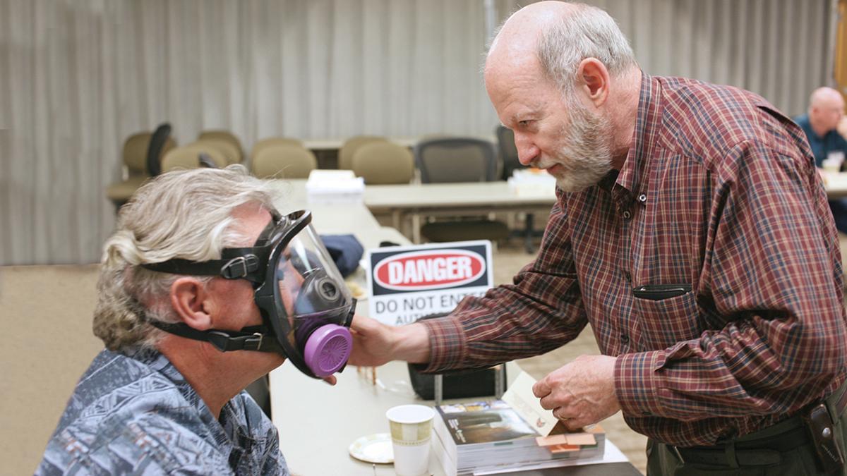 A man in a plaid shirt stands in a classroom helping a person adjust the respirator he is trying on.