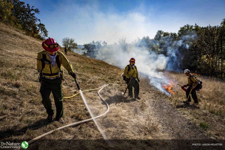 Three people in hard hats work on a hillside with a small fire burning, one spraying water from a hose.