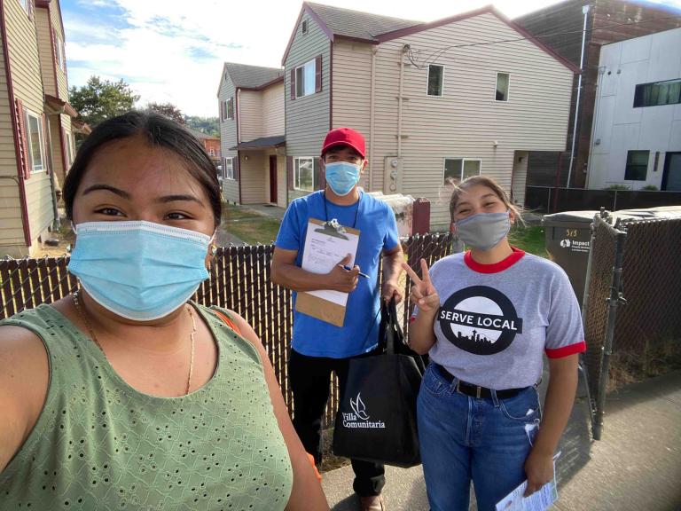 Three people with face masks on stand in front of a fence with houses in background. Person in middle holds a clipboard and pen, person on right gives a peace sign and is wearing a t-shirt that reads "SERVE LOCAL."