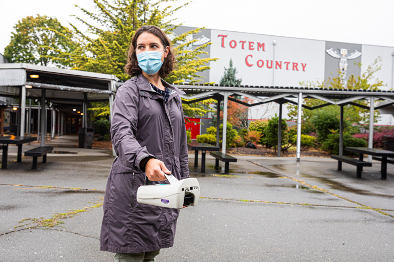EDGE member Elena Austin stands in a parking lot outside a school holding an air monitoring instrument.