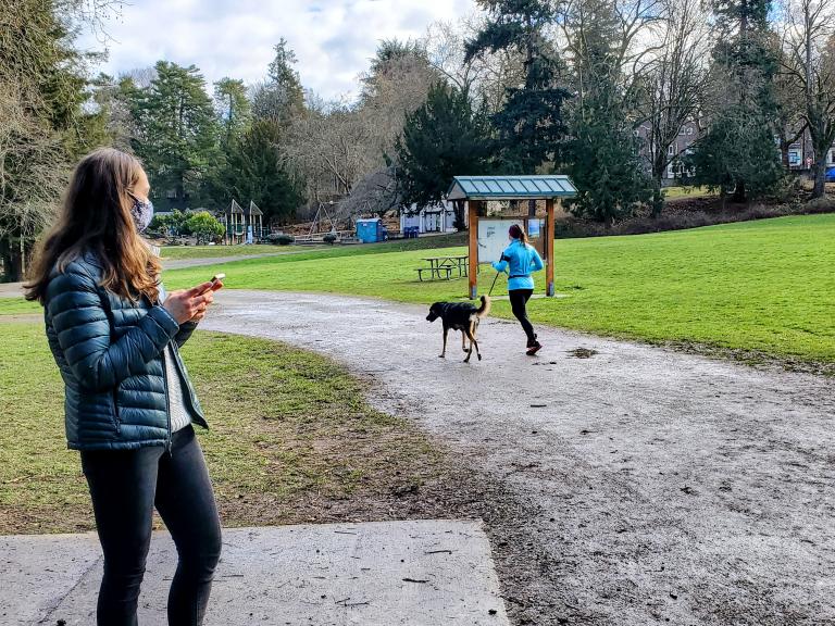 In a park, a woman in a face mask holds a mobile phone while observing a runner with a dog on a leash.
