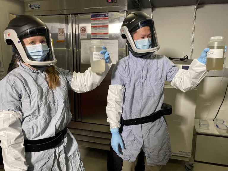 Two lab workers in protective suits and face shields hold up samples of wastewater in plastic bottles.