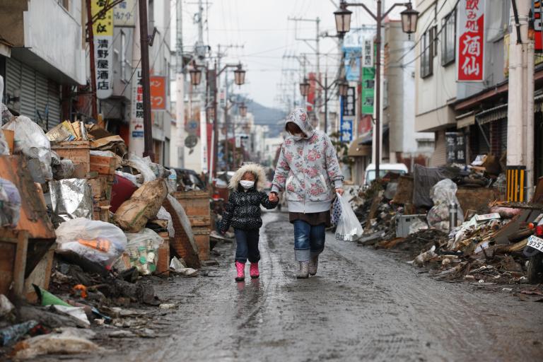 An adult and child walk hand in hand along a street in an urban area on a path cleared through debris. 