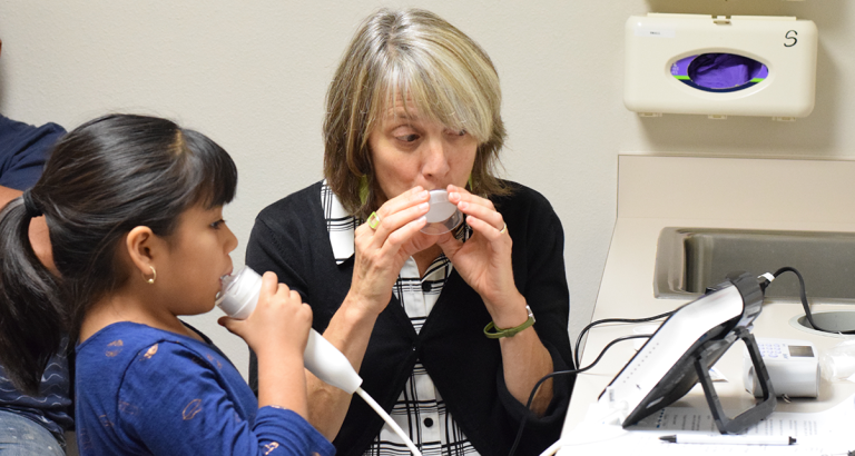 Dr. Catherine Carr demonstrates blowing into a filter while a child blows into a device measuring lung inflammation.