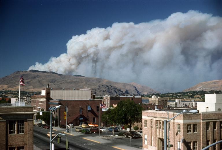 A plume of wildfire smoke behind the center of a small town showing brick buildings and cars. 