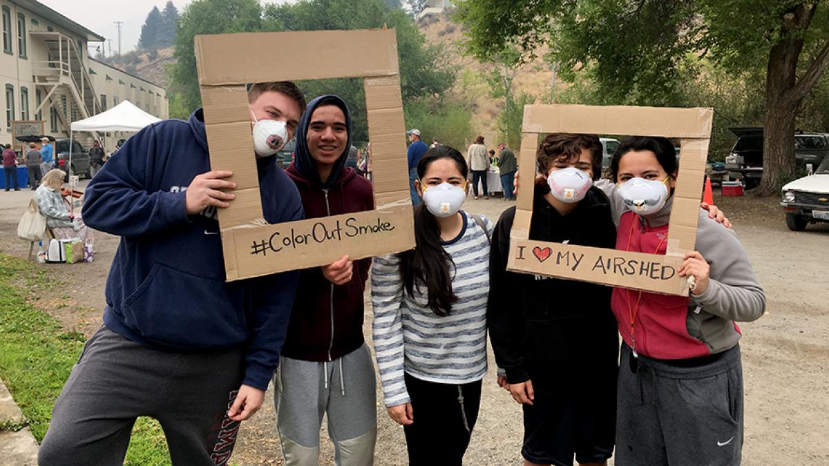 Teens pose in smoky air wearing masks and holding signs that say "I love my airshed" and "color out smoke"
