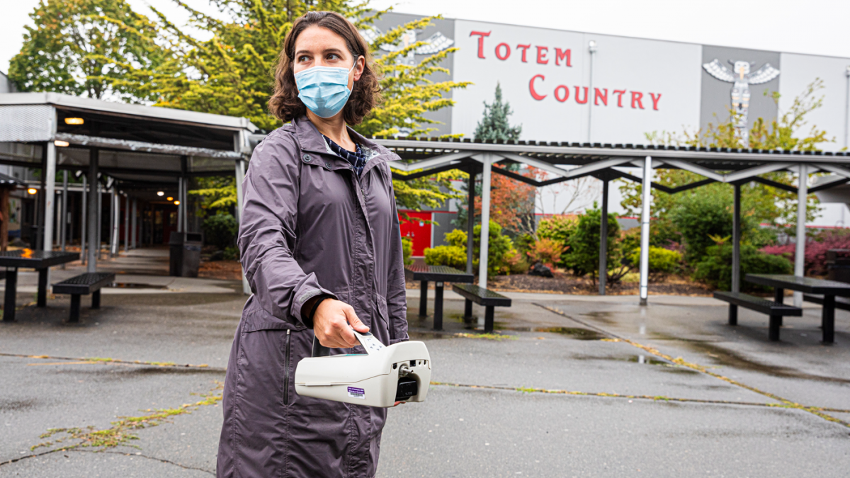 EDGE member Elena Austin stands in a parking lot outside a school holding an air monitoring instrument.