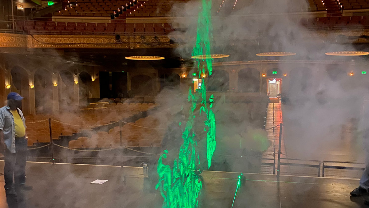 A neon green laser light highlights the airflow on the stage of a large theatre space.