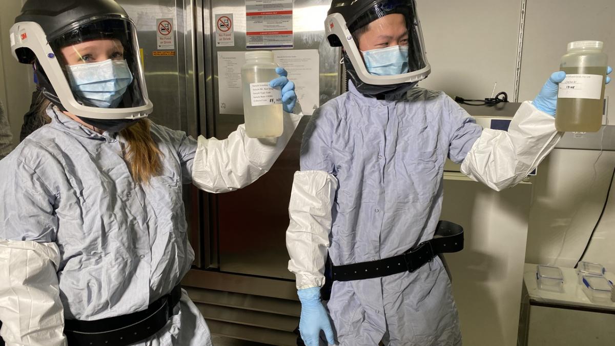 Two lab workers in protective suits and face shields hold up samples of wastewater in plastic bottles.