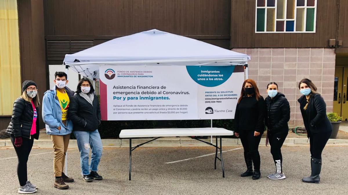 Six people in masks stand around a sign in Spanish offering financial assistance during the pandemic to immigrants. 