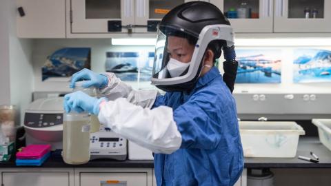 DEOHS Research Scientist Angelo Ong, in protective face shield helmet in a lab, holds two bottles of wastewater for analysis.