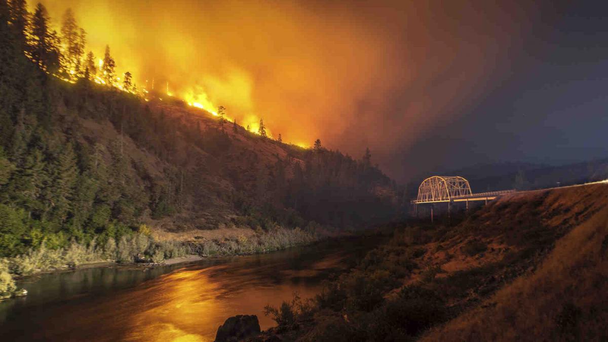A forest fire on a ridge against a darkened sky next to a river with a bridge crossing.