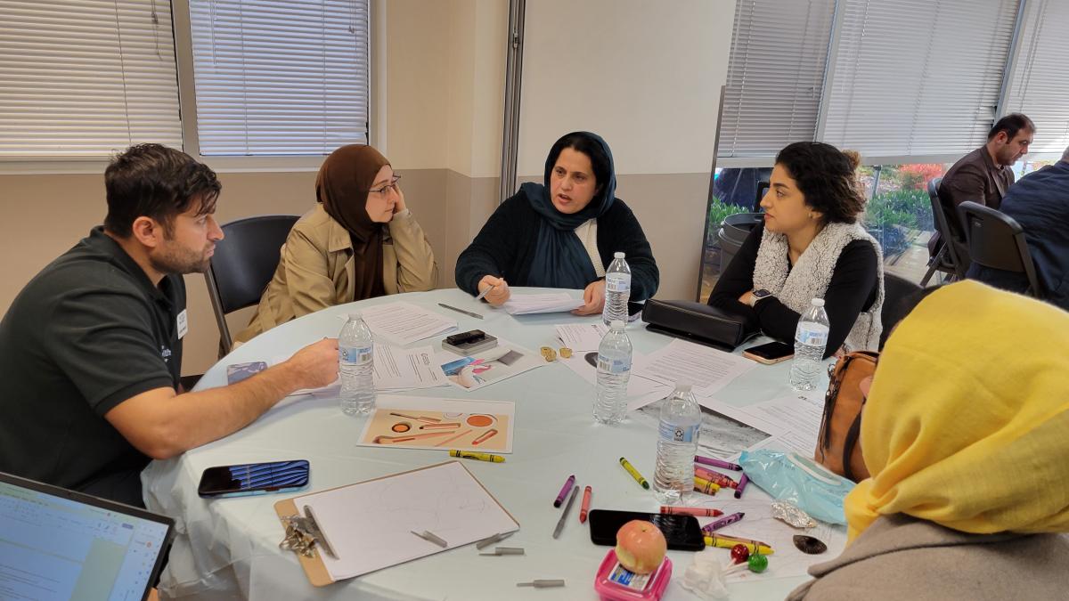 Five people sit around a round table with a white tablecloth with papers, crayons, plastic water bottles. One person in a headscarf is speaking and the others are listening.