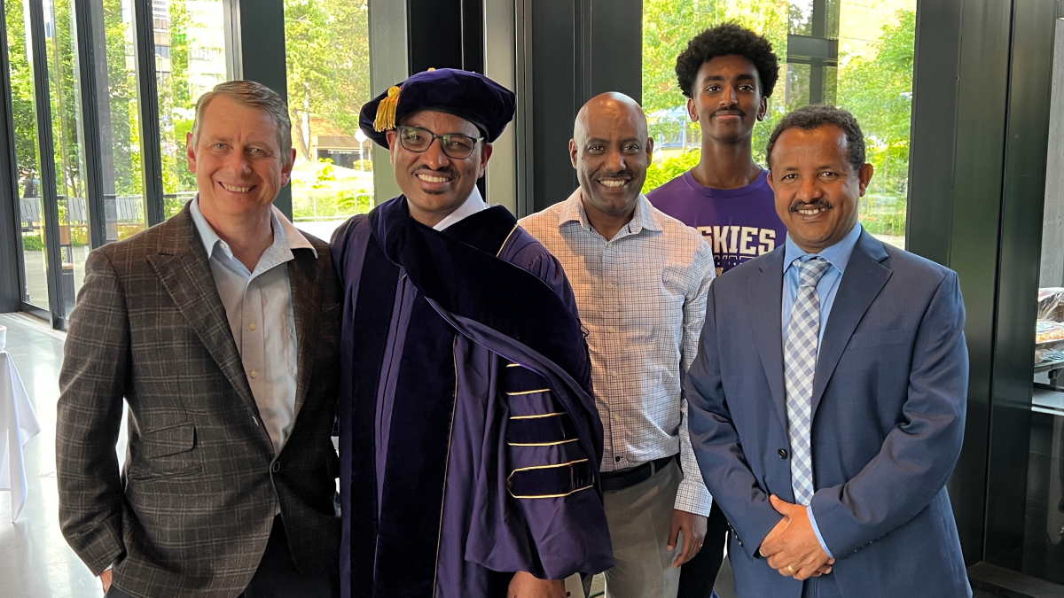 Five people stand together smiling. The second from left is wearing graduate regalia.