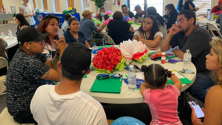 A table of seven farmworkers and a child sit in the foreground having a discussion, with other tables of people in the background. 