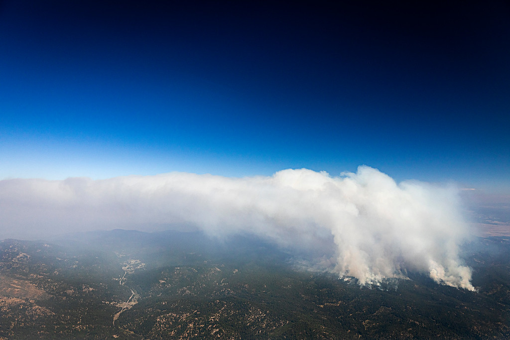 Wildfire smoke rises in plumes above a mountainous landscape in an aerial photo.