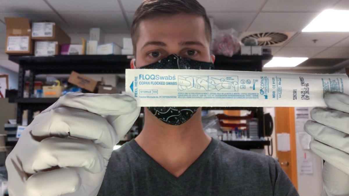 Man wearing a face mask and medical gloves stands in a lab holding a packaged swab in front of his face.