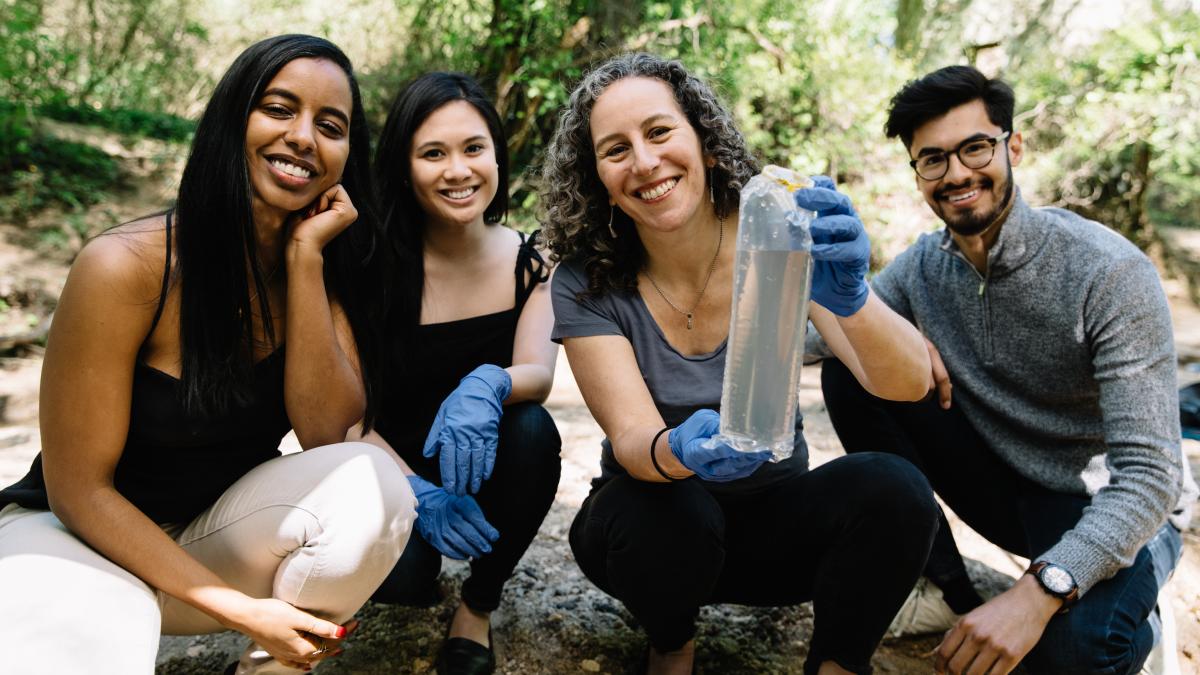 Karen Levy and three students crouch down outside with Levy holding a water sample in a plastic bag. 