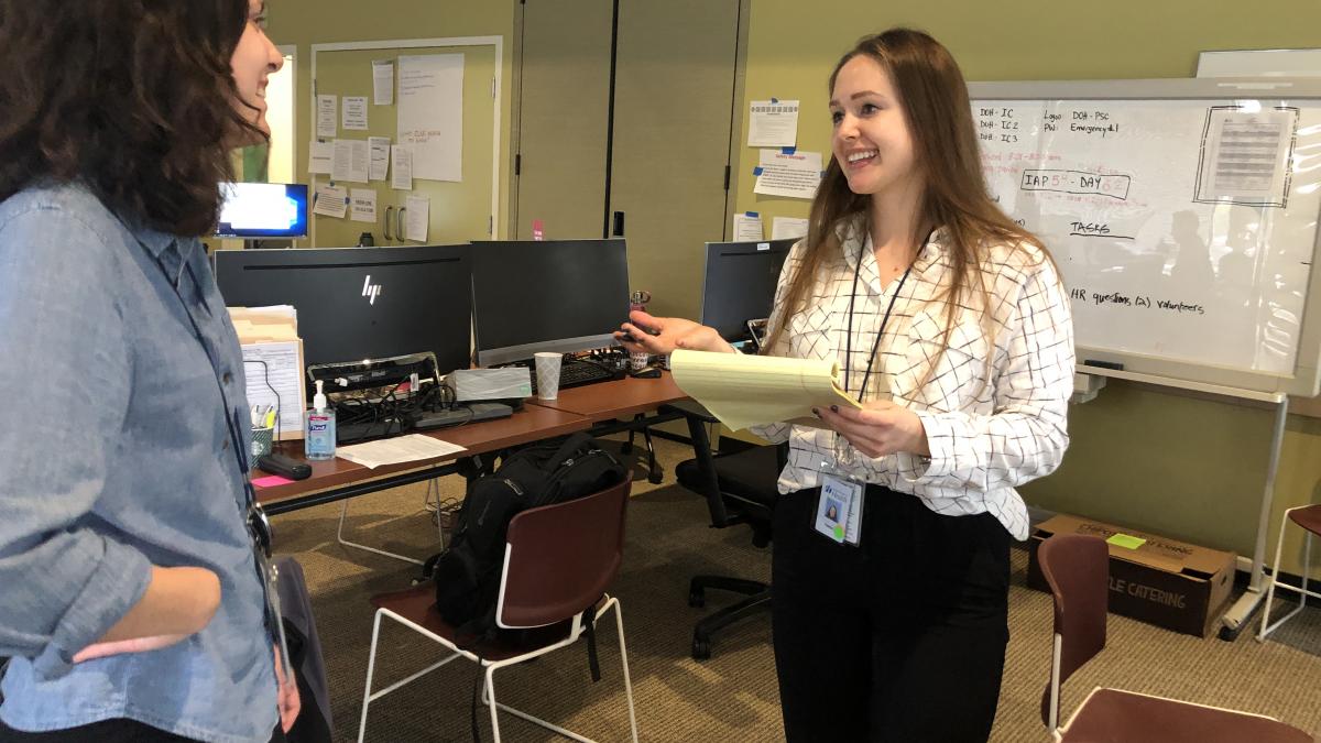 A woman in a white shirt talks with another woman in a blue shirt while standing in front of desks with computers.