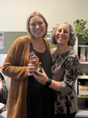 Becky Kann and Karen Levy stand together smiling and holding a trophy of a smiling poop emoji. 