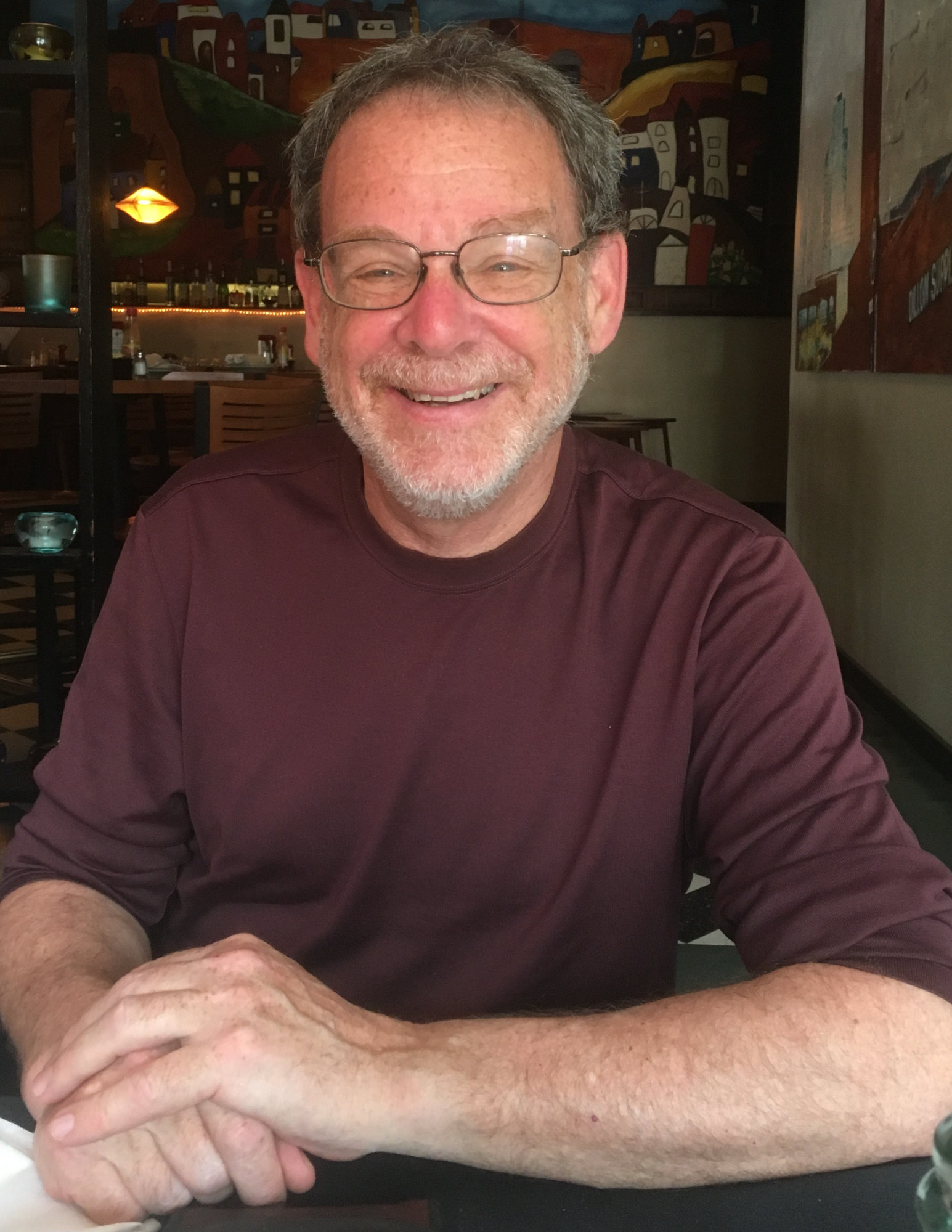 Robert Harrison wears long sleeved shirt, sleeves rolled up, and sits with hands folded, smiling at the camera
