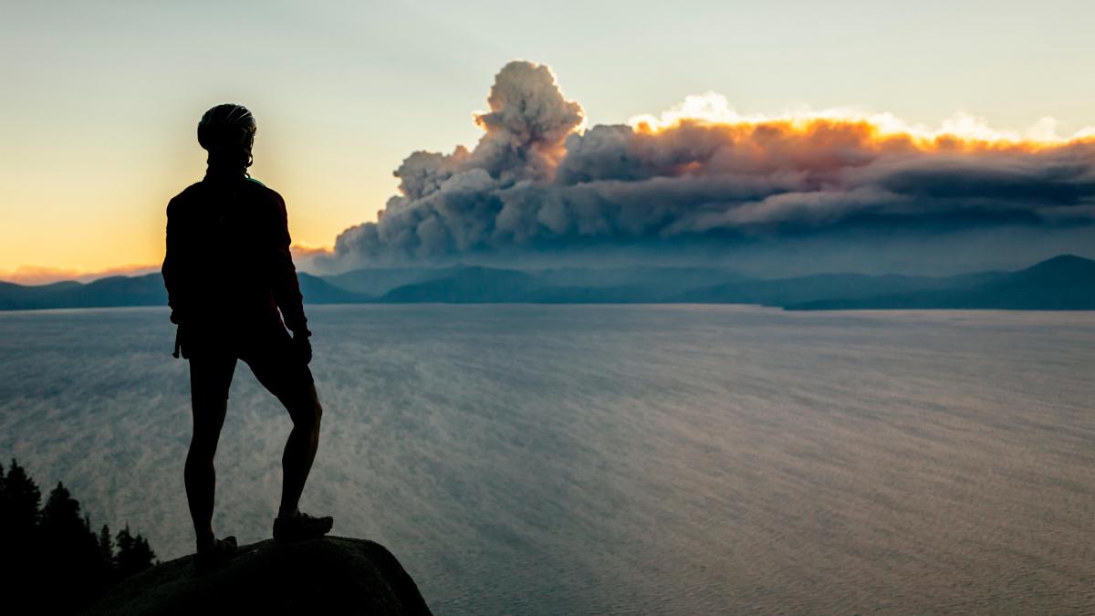 A woman in a bike helmet looks out over a lake toward wildfire smoke in the distance