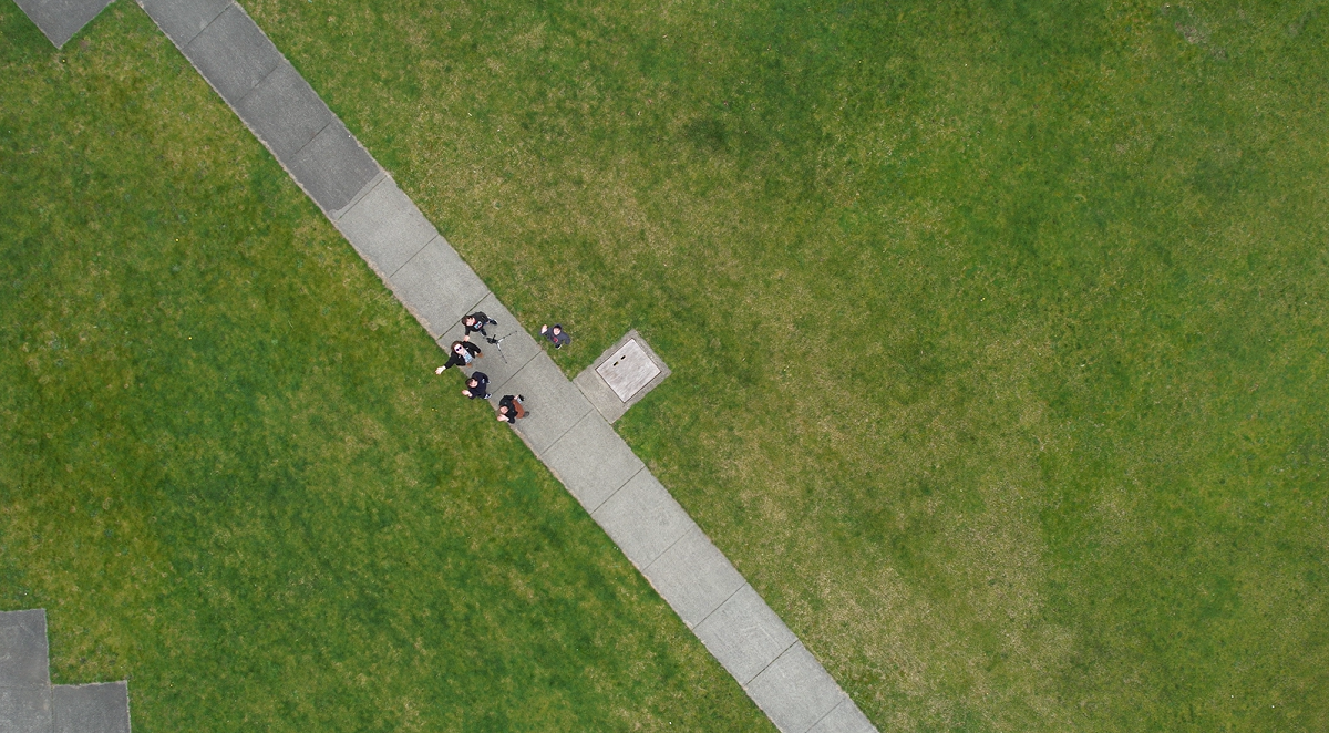 Aerial photo of four people waving from the ground on a sidewalk surrounded by grass.
