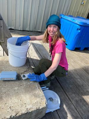 A woman in a blue cap kneels near a bucket on a dock with scientific instruments in her hands.