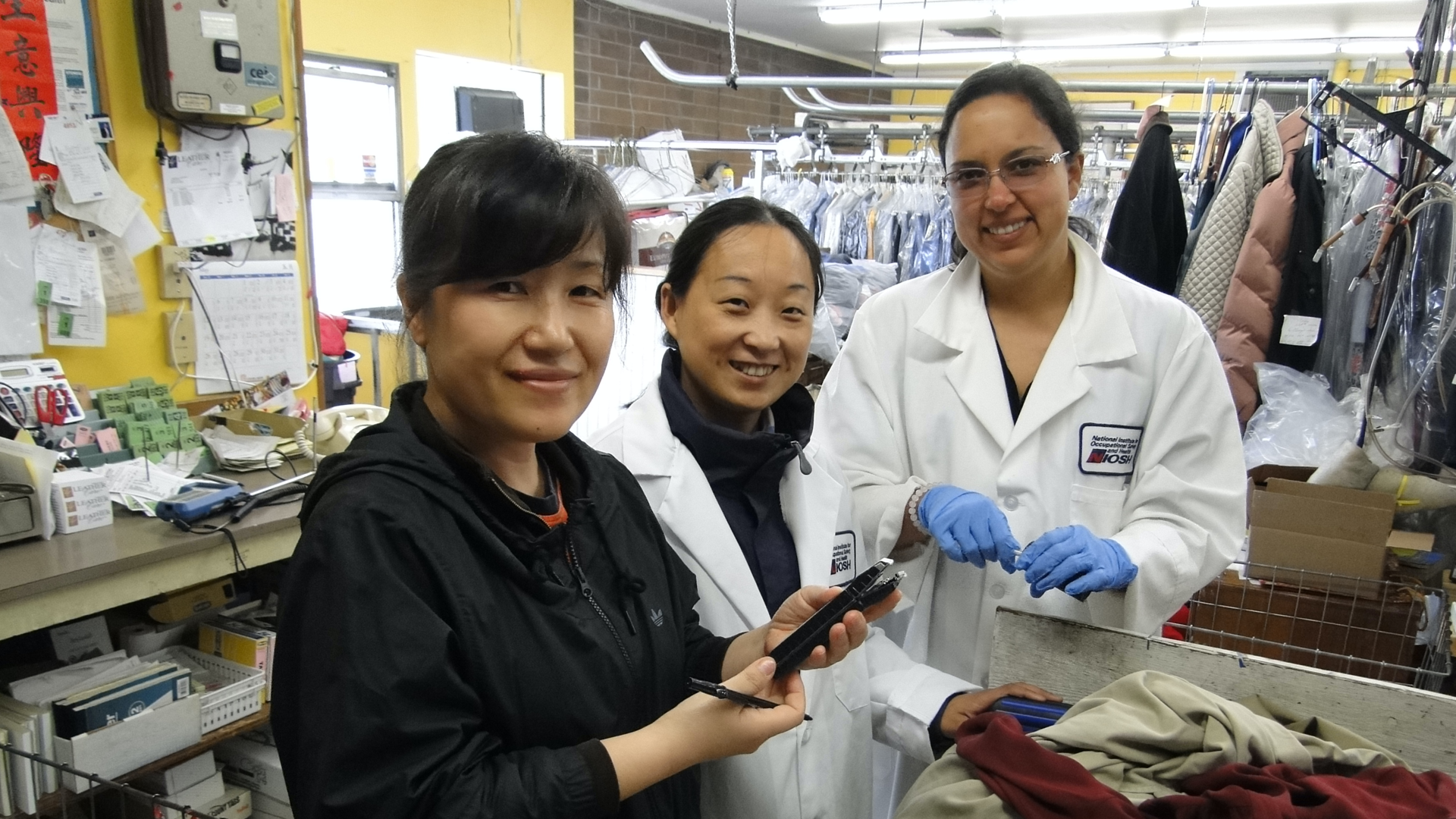 A woman in a white lab coat and glasses stands next to two other women in a dry-cleaning business.