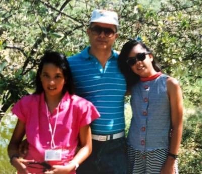 Twin girls stand outside under a tree with their father, who wears a white cap and blue shirt.