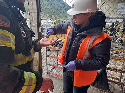 A woman in a hard hat and safety vest swabs a firefighter's hand.