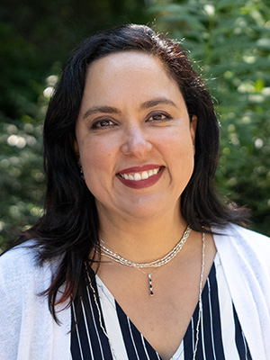 Headshot of Diana Ceballos smiling outside with shoulder length black hair, wearing a silver necklace with a blue and silver pendant, a striped navy blue and white shirt and a white cardigan sweater.