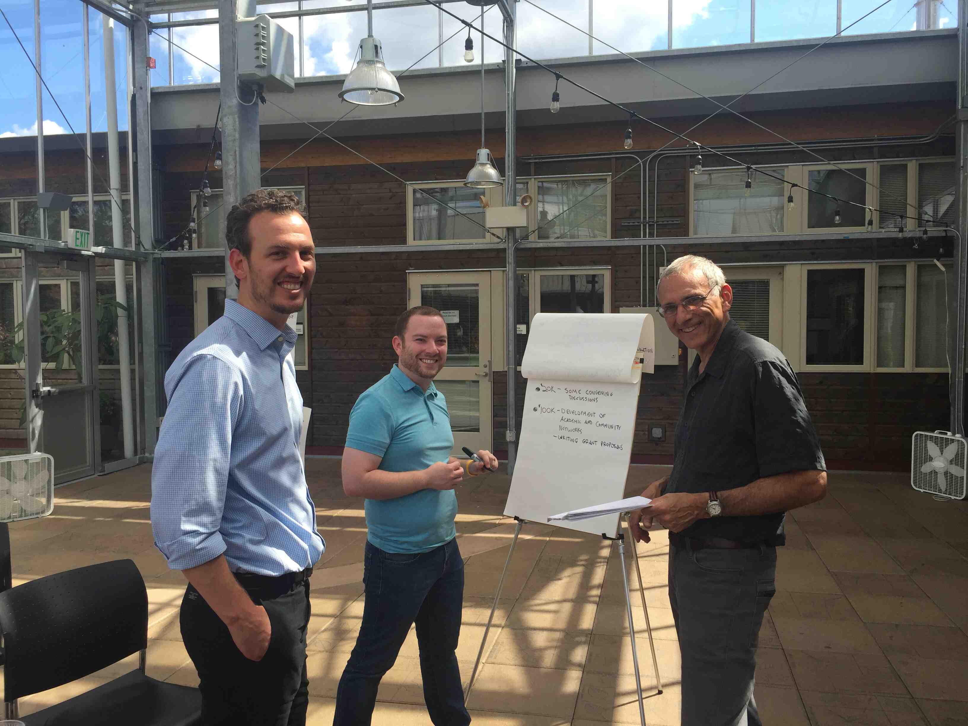 Three men smile at the camera while working on a flipchart.