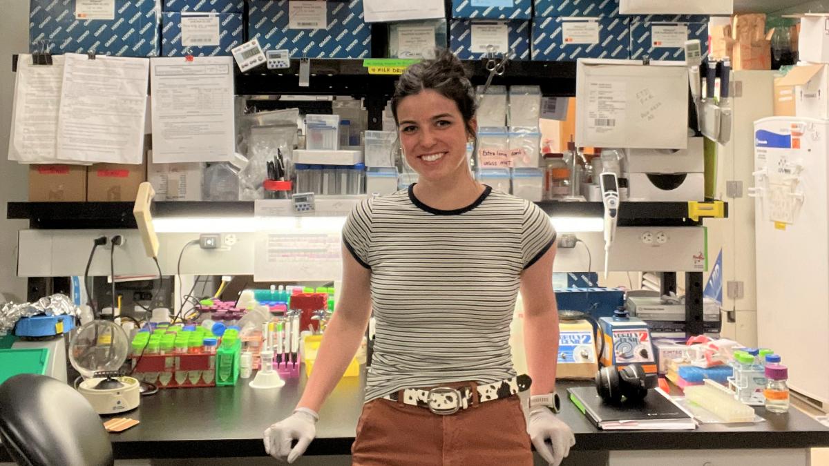 A woman in a striped T-shirt and wearing surgical gloves stands near a desk in a laboratory.